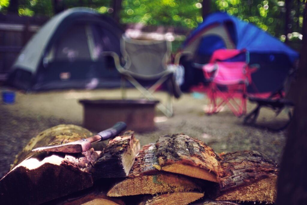 Camping for Beginners: How to Plan, Pack, and Arrive Without Stress Close-up of split firewood and an axe at a campsite with tents and chairs blurred in the background.